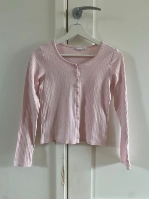 Ljusrosa ribbad kofta med knappar - Söt ljusrosa ribbad kofta med långa ärmar och rund halsringning. Koftan har knäppning framtill med matchande rosa knappar och är tillverkad i ett mjukt, stretchigt material. Perfekt att styla över ett linne eller t-shirt.