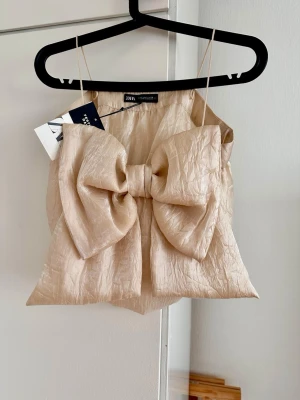 Beige bow top ftom Zara - Supersnygg beige topp från Zara med tunna axelband och en stor rosett framtill. Materialet har en glansig satinlook och lätt skrynklig textur. Perfekt för dig som vill sticka ut med en unik och trendig topp.