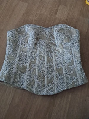 Vit spetskorsett med blommönster - Supersnygg vit korsett med detaljerat blommigt spetsmönster och transparent mesh. Axelbandslös modell med markerade kupor för extra form. Perfekt att styla till jeans eller kjol för en trendig look.