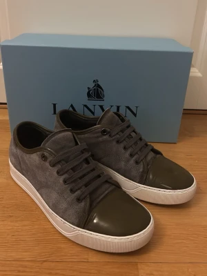 Lanvin captoe DBB1 - Snygga sneakers från Lanvin i grå mocka med mörkgrå lackad tå och matchande snörning. Skorna har en vit sula som ger en clean kontrast och en klassisk låg silhuett. Perfekta för dig som gillar stilrena detaljer och exklusiva material. Storlek 41~. Sulan är reparerad med skokit från shoehero. PRISET KAN FÖRHANDLAS