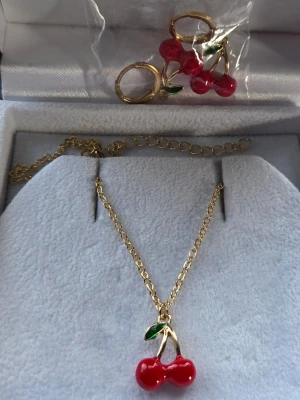 Guldigt halsband med körsbärshänge - Säljer ett guldigt halsband med ett sött körsbärshänge i rött och grönt. Hänget har två röda pärlor som körsbär och ett litet grönt blad. Perfekt för dig som gillar färgglada och lekfulla accessoarer. Matchande örhängen finns också med samma design. 45 cm 