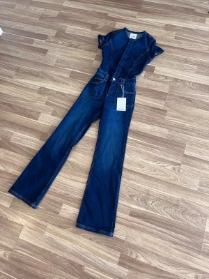 Jeansoverall - Snygg mörkblå jeansjumpsuit från Pull&Bear med korta ärmar och raka ben. Jumpsuiten har markerade sömmar, rund halsringning och fickor fram. Perfekt för dig som vill ha en cool och trendig look i denim.