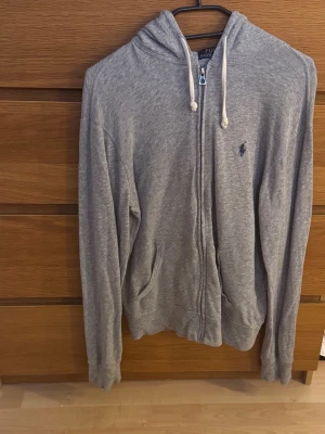 Grå zip hoodie från Polo Ralph Lauren - Snygg hoodie från Polo Ralph Lauren med dragkedja framtill, två fickor och justerbar huva med vita snören. Klassisk logga broderad på bröstet. Tillverkad i mjukt och bekvämt material, perfekt för en avslappnad stil.