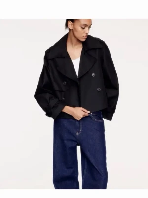 Svart kort trenchcoat från H&M - Stilren svart trenchcoat från H&M med dubbelknäppning och breda slag. Jackan är kortare i modellen och har justerbara ärmslut med knappar. Perfekt för att ge en cool och modern look. Materialet är lätt och passar bra till jeans eller dressade byxor. Storlek M och perfekt för dig som bär S och vill ha lite lösare passform!