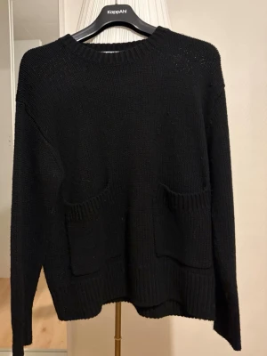Svart stickad tröja från Zara - Snygg svart stickad tröja från Zara med två stora fickor framtill. Tröjan har rund halsringning, långa ärmar och ribbade muddar vid ärmslut och nederkant. Perfekt för lager på lager och enkel att matcha med olika outfits.