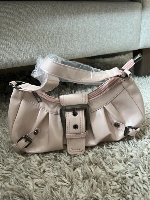 Ljusrosa axelväska med spänne - Trendig ljusrosa handväska i glansigt konstläder med bred axelrem och stora silvriga spännen framtill. Väskan har veckade detaljer och dragkedja upptill. Perfekt för dig som gillar en söt och stilren look. Ej äkta läder (plast-ish material). Notera att dragkedjan kan vara lite trög.