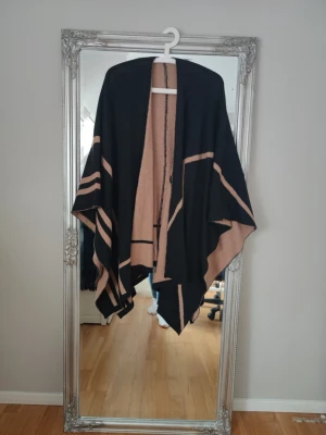 Svart och beige sjal med grafiskt mönster - Stilren poncho i svart och beige med grafiskt mönster och öppet fall. Sjalen har breda kanter och asymmetrisk form, vilket ger en cool och avslappnad look. Perfekt att slänga över axlarna för extra värme och stil. Materialet känns mjukt och följsamt.
