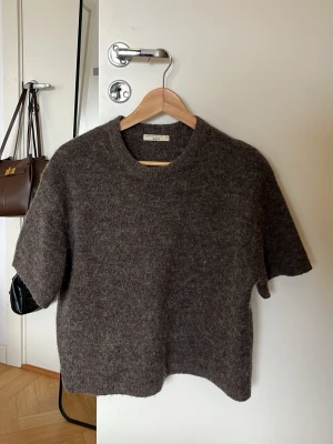 Mörkbrun stickad t-shirt Gina Tricot - Kortärmad stickad t-shirt från Gina Tricot i mörkbrunt, tillverkad i ett mjukt och fluffigt material. Rund halsringning och rak passform ger en avslappnad look. Perfekt att styla lager på lager eller bära som den är.