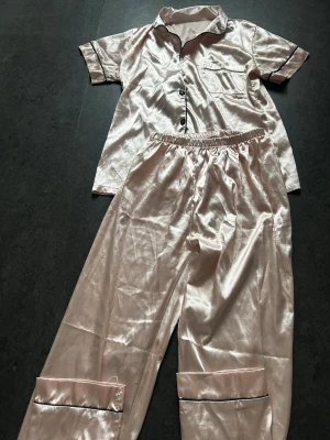 Ljusrosa satinpyjamas med svarta detaljer - Snygg ljusrosa pyjamas i glansigt satinmaterial med kortärmad skjorta och långa byxor. Skjortan har svarta knappar, bröstficka och svarta passpoaler längs krage och ärmslut. Byxorna har resår i midjan och matchande svarta detaljer vid benslut. Perfekt för mysiga kvällar.