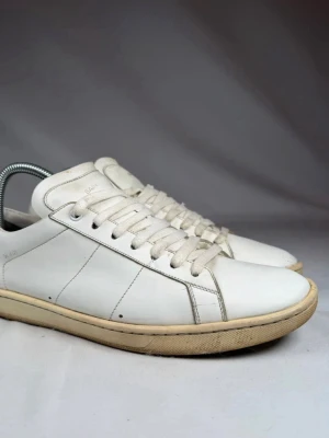 Vita Saint Laurent  - Klassiska vita sneakers från Saint Laurent i slätt läder med vita snören och diskret logga på plösen. Snygg minimalistisk design med lågt skaft och beige sula som ger en clean look. Perfekta för dig som gillar stilrena och tidlösa skor.