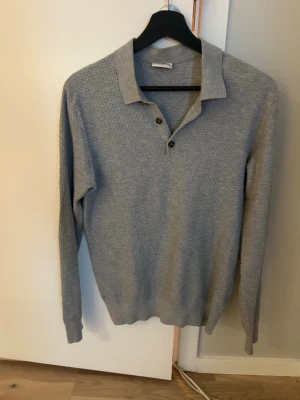 John Henric stickad/kashmir tröja - Snygg ljusgrå stickad tröja med krage och två knappar framtill. Tröjan har lång ärm och ribbade muddar vid ärmslut och nederkant. Liten i storlek. Inköpt på Johan Henric i Sturegallerian i Stockholm