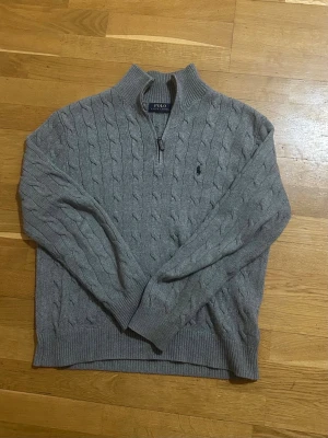 Grå kabelstickad tröja Polo Ralph Lauren - Säljer denna snygg gråa kabelstickade tröjan från Polo Ralph Lauren med 1/2 zip. Tröjan har långa ärmar, ribbade muddar och den klassiska broderade loggan på bröstet. Perfekt för dig som gillar stilrena och tidlösa plagg.