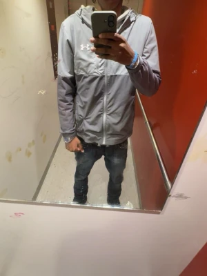 Grå Under Armour vindjacka med huva - Snygg grå vindjacka från Under Armour med huva och dragkedja framtill. Jackan har en sportig design med logotyp på bröstet och är tillverkad i ett lätt material som passar perfekt för aktiva dagar. Jackan har långa ärmar och en modern passform.