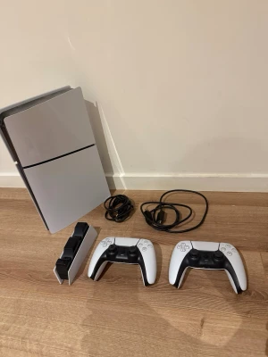 PlayStation 5 med två handkontroller - PlayStation 5 konsol i vit och svart design från Sony. Medföljer två trådlösa DualSense-kontroller, laddstation och kablar. Konsolen har en futuristisk look med rundade kanter och matta ytor. Perfekt för gaming och multiplayer-sessioner. Ena kontrollen har lite stick drift men inget som märks tydligt av. Går att förhandla 