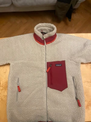 Beige teddyjacka från Patagonia - Säljer en beige teddyjacka från Patagonia med dragkedja framtill, hög krage och kontrasterande vinröda detaljer. Jackan har två sidofickor med dragkedja och en stor bröstficka i vinrött tyg med logga. Materialet är mjukt och fluffigt, perfekt för kalla dagar.