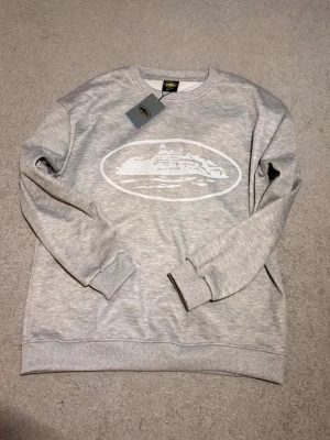 Corteiz sweatshirt grå - Snygg grå sweatshirt med ett vitt tryck av en ö eller landskap på bröstet. Tröjan har rund hals, ribbade muddar vid ärmslut och nederkant. Perfekt för en avslappnad stil och passar till det mesta. Materialet känns mjukt och bekvämt.