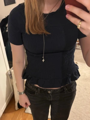 Mörkblå topp med smock från Gina Tricot - Säljer en mörkblå topp från Gina Tricot med smockad front och baksida samt volangkant nedtill. Toppen har korta ärmar och rund halsringning. Perfekt för en stilren och trendig look.