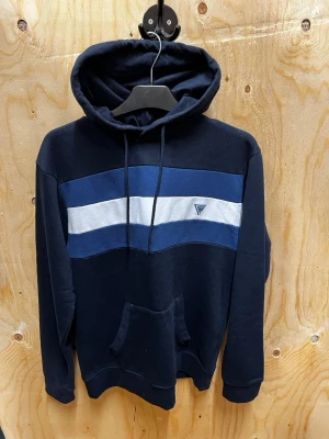 Stilren mörkblå hoodie - Stilren mörkblå hoodie med breda blå och vita ränder över bröstet och en liten logga på vänster sida. Perfekt för en avslappnad och sportig stil.