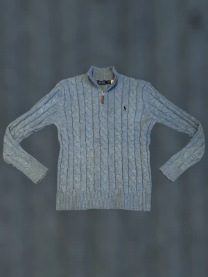 Helt ny! Polo Ralph Lauren 1/4 zip - Kabelstickad grå tröja från Polo Ralph Lauren med halv dragkedja och hög krage. Tröjan har ribbade muddar och nederkant samt den klassiska broderade loggan på bröstet. Perfekt för lager på lager och snygg till jeans.