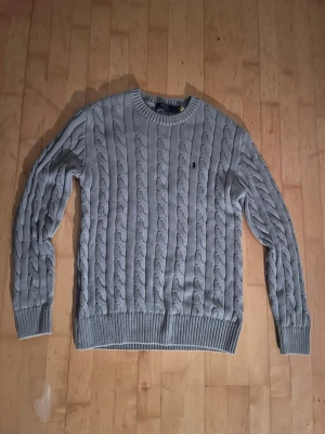 Ralph Lauren - Snygg grå kabelstickad tröja från Polo Ralph Lauren. Tröjan har rund halsringning, ribbade muddar och nederkant samt en liten broderad logga på bröstet. Perfekt för lager på lager och en klassisk look. Storlek M, pris kan diskuteras.