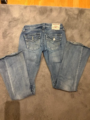True Religion ljusblå bootcut jeans - Säljer ett par mörkblå jeans från True Religion med bootcut-modell. Jeansen har snygga bakfickor med knappar. Väldigt bra skick och låg midjade