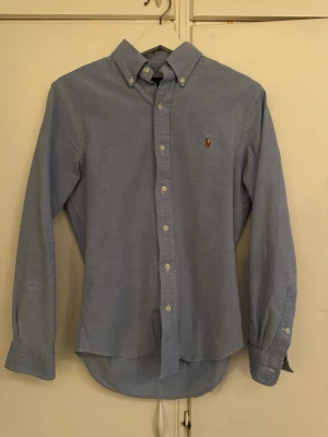 Blå skjorta från Ralph Lauren - Klassisk ljusblå skjorta från Ralph Lauren med button-down krage och broderad logga på bröstet. Skjortan har långa ärmar, vita knappar och är tillverkad i mjuk bomull. Perfekt till både jeans och chinos för en clean look.