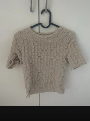 blus  - Trendig beige croppad topp med ribbad struktur och korta ärmar. Perfekt till höga jeans eller kjol för en avslappnad men snygg look. Materialet känns mjukt och följsamt mot huden. använd en gång