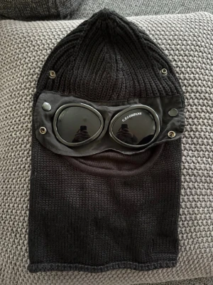 Svart skidmask från C.P. Company - Säljer en svart stickad balaclava från C.P. Company med inbyggda goggles och detaljer i metall. Mössan har en unik design med glasögon integrerade i tyget och är tillverkad i mjukt, värmande material. Perfekt för dig som vill sticka ut med streetwear-vibe.