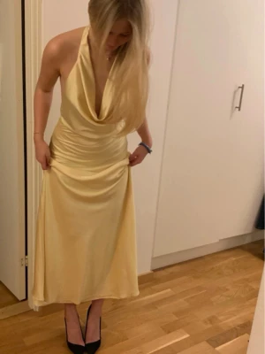 Balklänning  - Elegant långklänning i satin med halterneck och djup urringning framtill. Klänningen har öppen rygg och knyts i nacken för en snygg siluett. Perfekt för dig som vill sticka ut med en lyxig och stilren look. 