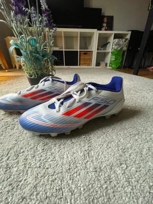 Adidas vita och blå fotbollsskor - Säljer ett par Adidas fotbollsskor i vitt med blå och röda detaljer. Skorna har snörning, låg profil och en ovandel i syntetmaterial med texten 'FIBERSKIN'. Insidan är blå och yttersulan är anpassad för spel på gräs. Perfekta för dig som vill sticka ut på planen. 88kr frakt betalas av köp.Är ute för förhandling