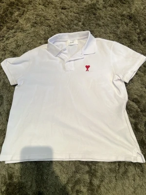 Vit pikétröja från Ami med rött hjärta - Klassisk vit pikétröja från Ami med korta ärmar och krage. På bröstet finns Ami:s ikoniska röda hjärtemblem. Tröjan är tillverkad i mjuk bomull och har knappar vid halsen för en clean look.