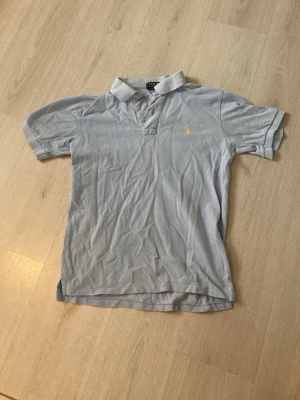Ljusblå piké tröja Ralph Lauren - Ljusblå piké t-shirt från Ralph Lauren med klassisk krage, korta ärmar och två knappar vid halsen. Liten gul broderad logga på bröstet. Perfekt för sommaren och kommande varma dagar. (Priset är ej hugget i sten). 