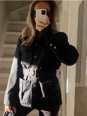 Svart jacka med bälte - Stilren svart jacka med markerat midjebälte och stora fickor framtill. Klassisk skjortkrage och knäppning med metallknappar. Perfekt för lager-på-lager och ger en cool, avslappnad look. Materialet ser ut att vara kraftig bomull eller bomullsblandning.