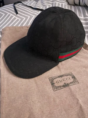 Svart Gucci keps med GG-mönster - Säljer en svart keps från Gucci med det klassiska GG-monogrammet över hela kepsen. På sidan finns en grön och röd rand som ger den en ikonisk look. Kepsen är tillverkad i canvasmaterial och har en böjd skärm. Perfekt för dig som vill ha en stilren och lyxig accessoar.