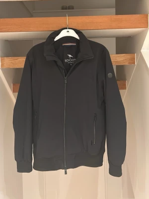 Svart vindjacka från Snoot - Stilren svart vindjacka från Snoot med hög krage och hel dragkedja framtill. Jackan har två sidofickor med dragkedja, ribbade muddar vid ärmslut och nederkant samt en diskret logotyp på ärmen. Perfekt för blåsiga dagar. 2500kr i ny pris