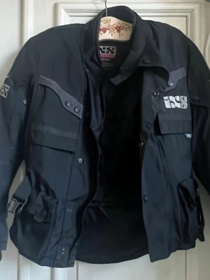 Svart motorcykeljacka från iXS - Säljer en svart motorcykeljacka från iXS med flera fickor, justerbara spännen i midjan och tryckta loggor på bröstet och ryggen. Jackan har hög krage, långa ärmar och stängs med dragkedja och knappar. 