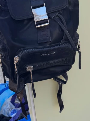 Svart ryggsäck från Steve Madden - Snygg svart ryggsäck från Steve Madden med flera dragkedjefack och silverfärgade detaljer. Tillverkad i ett glansigt tygmaterial med justerbara axelband och en stor metallspänne framtill. Perfekt för dig som vill ha en trendig och praktisk väska.nyprs1199kr säljes 249kr