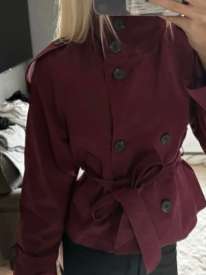 Vinröd trenchcoat med knytskärp - Stilren vinröd trenchcoat med stora svarta knappar och bälte i midjan. Jackan har axelklaffar, bred krage och dubbelknäppning. Perfekt för dig som vill ha en klassisk men trendig look. Materialet ser ut att vara bomull eller polyester.