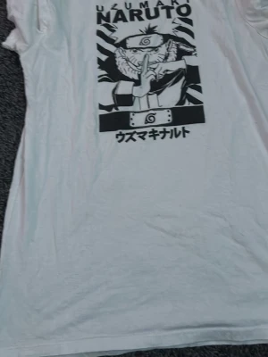 Vit Naruto t-shirt med tryck - Säljer en vit t-shirt med ett stort svartvitt tryck av Uzumaki Naruto från anime-serien. T-shirten har korta ärmar och rund hals. Perfekt för dig som älskar manga och vill ha en unik och snygg tee i garderoben.