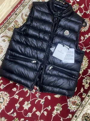 Svart dunväst från Moncler - Snygg svart dunväst från Moncler med glansig finish och klassisk logga på bröstet. Västen har två dragkedjeförsedda fickor framtill och hög krage. Perfekt för kalla dagar när du vill hålla stilen. Materialet är dunfyllning och yttertyget är blankt.