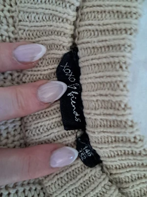 Beige stickad tröja XOXO Friends - Snygg beige stickad tröja från XOXO Friends med rund halsringning och ribbade muddar. Tröjan har ett klassiskt, lite kortare snitt och är gjord i ett mjukt, grovstickat material. Perfekt för lager på lager och enkel att matcha.