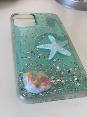 Turkost mobilskal med glitter och sjöstjärna - Säljer ett turkost mobilskal dekorerat med silverglitter, små stjärnor och en 3D-sjöstjärna samt snäcka fylld med färgglada pärlor. Skalets material är plast och det har en genomskinlig finish med havsinspirerad design. Perfekt för dig som vill ha ett unikt och färgglatt skal.