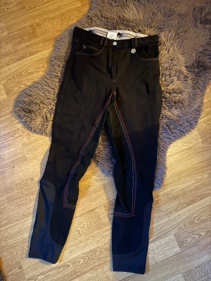 Svarta ridbyxor från Copperfield - Snygga svarta ridbyxor från Copperfield med röda kontrastsömmar och detaljer i mockaimitation på insidan av benen. Byxorna har klassisk femficksdesign och logga på knappen. Perfekta för dig som gillar hästar och vill ha en sportig look.