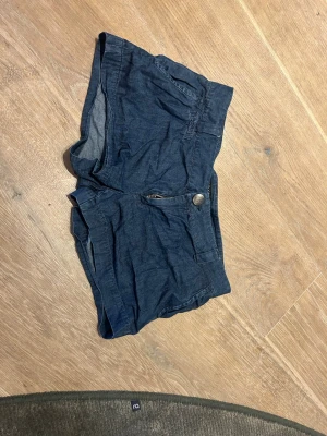 Mörkblå shorts med dragkedja och lådmidjade  - Säljer ett par mörkblå shorts med dragkedja och knapp framtill och lågmidjade. Shortsen har fickor både fram och bak och är tillverkade i ett mjukt bomullsmaterial. Perfekta för varma dagar och enkel att matcha med olika toppar.