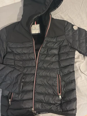 Svart dunjacka med huva från Moncler - Snygg svart dunjacka från Moncler med huva och dragkedja framtill. Jackan har två framfickor med dragkedja och Moncler-logga på ärmen. Detaljer i rött, vitt och blått längs dragkedjan ger en sportig touch. Perfekt för kalla vinterdagar.
