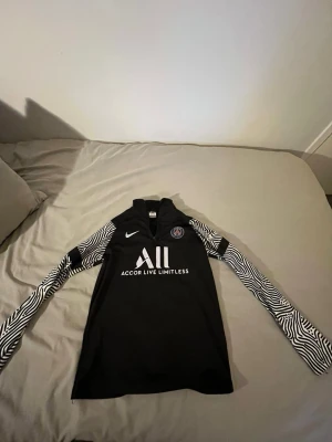 PSG träningströja svart/vit Nike - Snygg svart långärmad träningströja från PSG med vita zebramönstrade ärmar. Nike-logga och PSG-märke på bröstet samt texten 'ALL Accor Live Limitless' framtill. Perfekt för fotbollsträning eller gymmet.