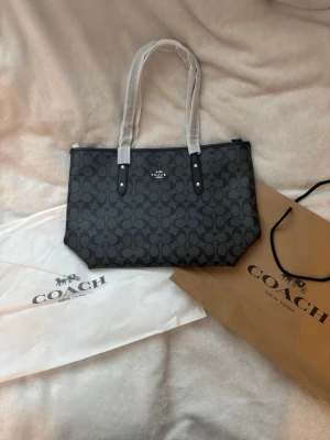Svart handväska från Coach - Snygg svart handväska från Coach med klassiskt monogrammönster och silverfärgad logga framtill. Väskan har två långa handtag och är tillverkad i tåligt material med detaljer i svart konstläder. Perfekt för dig som gillar stilrena accessoarer.