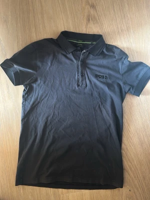 Svart pikétröja från BOSS - Klassisk svart pikétröja från BOSS med korta ärmar och krage. Tröjan har knappar framtill och diskret BOSS-logga broderad på bröstet. Tillverkad i mjuk bomull som känns skön mot huden. Perfekt till jeans eller shorts. Den ser kanske lite skrynklig ut, men de är bara och stryka den. Jag kan erbjuda det efter köp. Storlek S men funkar som M om man gillar tajtare look. Nypris 1200kr från boss butik på VÄLA. Säljer nu för 700 med original boss kartong till.