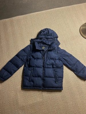 Mörkblå pufferjacka från Polo Ralph Lauren - Snygg mörkblå pufferjacka från Polo Ralph Lauren med broderad logga på bröstet. Jackan har huva, dragkedja framtill och två sidofickor med dragkedja. Perfekt för kalla vinterdagar och riktigt skön att bära. Materialet är slitstarkt och håller värmen bra.