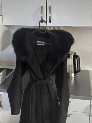 Svart kappa med fuskpäls - Stilren svart kappa med knytskärp i midjan och stor huva kantad med mjuk fuskpäls. Lång modell med markerad midja och raka ärmar. Perfekt för kalla dagar när du vill vara både varm och snygg.